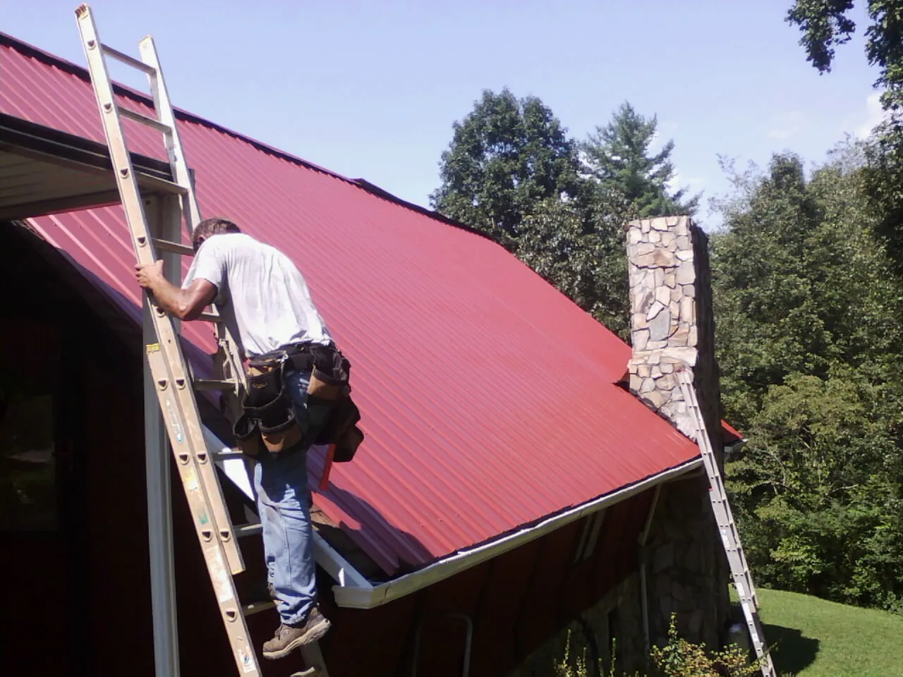 Roofer on a ladder beside a red metal roof and stone chimney — Soffit & Fascia Repair by Roofing Brothers in Kellerton